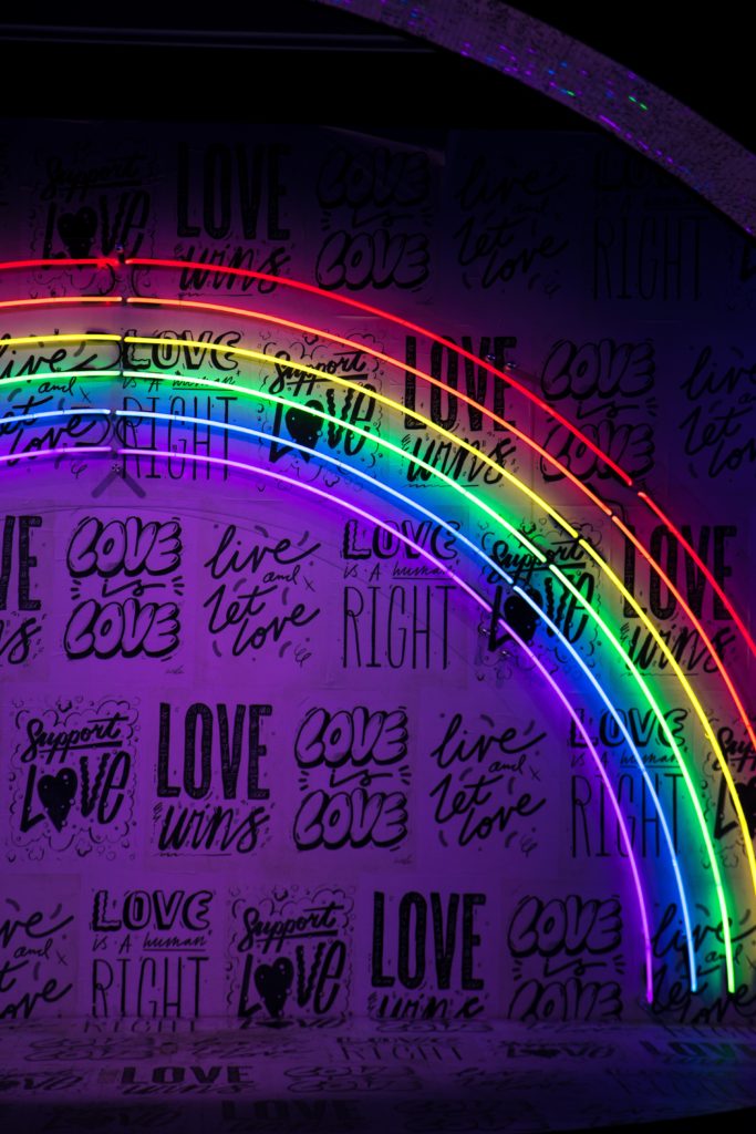 Rainbow neon lights, against a wall that has 'Love is Love' and 'Live and let Love' and other love slogans written on it.